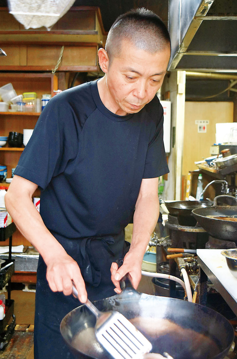 厨房（ちゅうぼう）で鍋を持つ店主の飯島義行さん＝本庄市前原の「味良久」