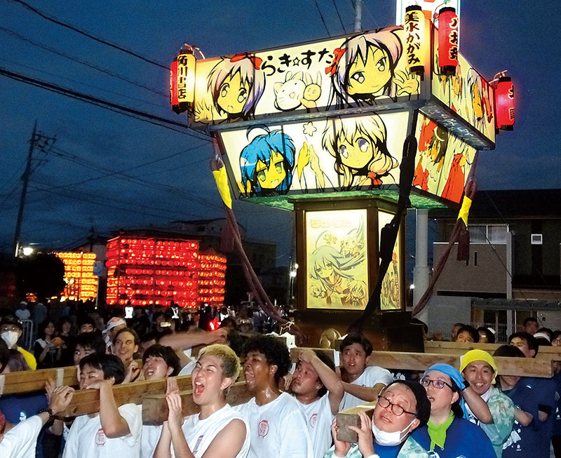 八坂祭で、新調されたらき☆すた神輿を担ぐファン。奥は地元町会の提燈山車＝28日夜、久喜市鷲宮