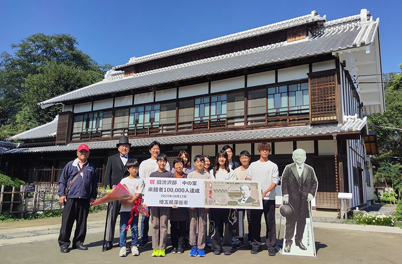 10万人目の来館者となった茨城県小美玉市の小松正二さん家族と深谷市の小島進市長（後列左から2人目）＝12日午前、深谷市血洗島の旧渋沢邸「中の家」